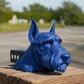 A blue ceramic dog head sculpture is displayed on a wooden post, with a blurred background of trees and a road.