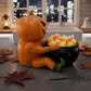 A Halloween-themed scene featuring a carved pumpkin sitting in a black pot filled with candy corn, surrounded by autumn leaves and a spider web.