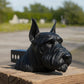 A black dog head sculpture is displayed on a wooden post, with a blurred background of trees and greenery.