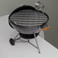 A small black and silver charcoal grill with a wire rack on top, sitting on a white surface.