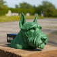 A green ceramic dog head sculpture is displayed on a wooden post, with a blurred background of trees and a road.