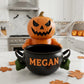 A Halloween-themed ceramic pumpkin with a carved face sits inside a black pot with the name &quot;Megan&quot; written on it, surrounded by autumn leaves on a kitchen counter.