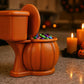 A Halloween-themed toilet-shaped candy holder filled with colorful candies sits on a kitchen counter, with two lit candles and carved jack-o-lanterns nearby.