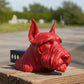 A red dog head sculpture is displayed on a wooden post, with a blurred background of trees and a road.
