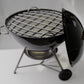 A small black grill with a metal grate and a wooden handle, placed on a white surface.