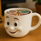 A white coffee mug with a smiling face design sits on a wooden surface, surrounded by coffee pods labeled with different types of coffee.