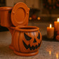 a Halloween-themed toilet with a carved pumpkin face, placed on a kitchen counter. In the background, there are two lit candles and two carved jack-o'-lanterns.