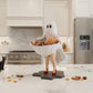 A Halloween-themed scene featuring a ghost figurine holding a bowl of candy, with a carved jack-o-lantern, string lights, and a kitchen counter in the background.