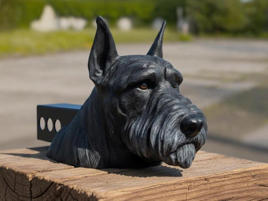 A black dog head sculpture is displayed on a wooden post, with a blurred background of trees and a road.