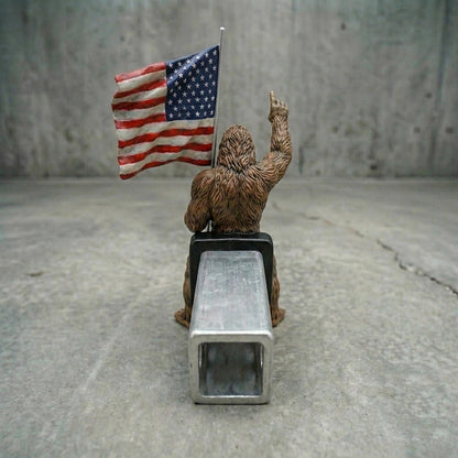 A toy figure of a Sasquatch holding an American flag stands on a metal object against a concrete background.