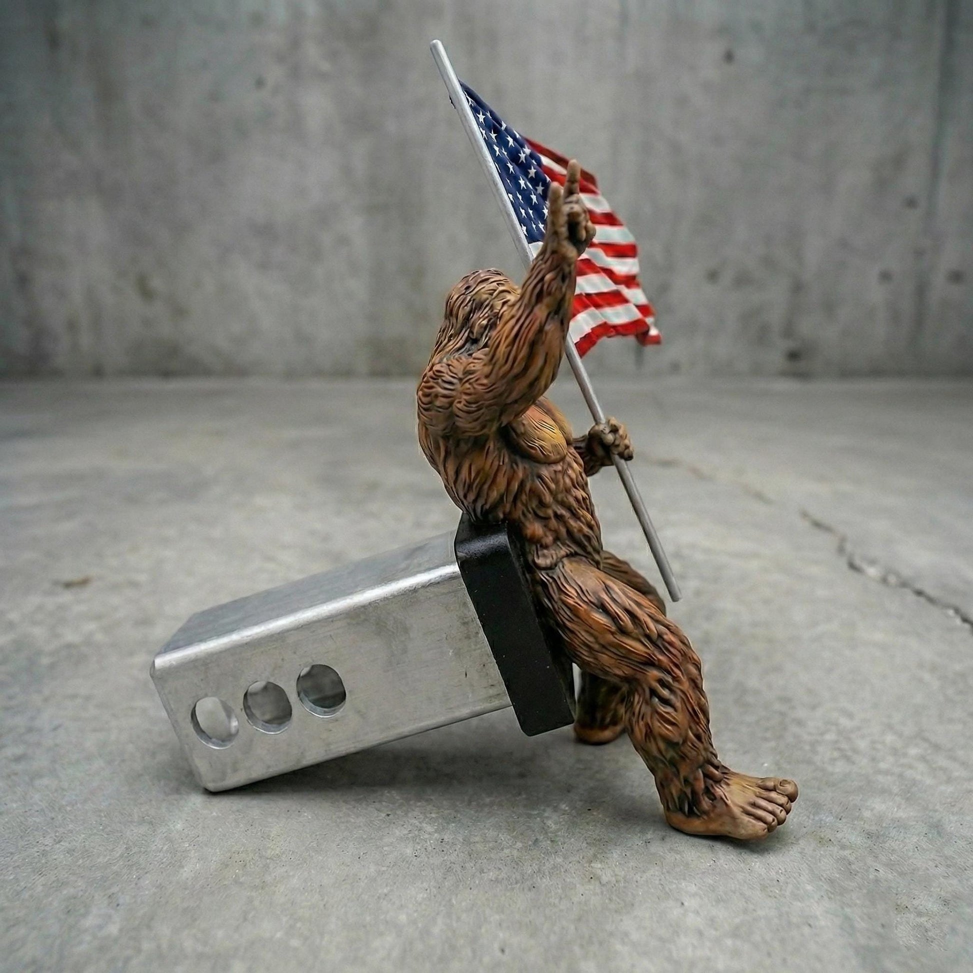 A brown, furry toy figure holding an American flag is sitting on a metal bumper attached to a concrete surface.